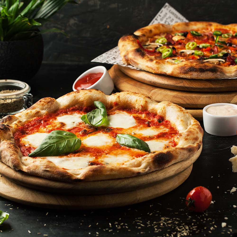 En sprød pizzabund med gylden kant er dækket af en rød tomatsauce og skiver af smeltet mozzarella. Friske basilikumblade er strategisk placeret ovenpå, hvilket tilfører farve og duft. I baggrunden ses en anden pizza med grøntsager og skinke, samt et par små skåle med sauces og krydderier. En cherry-tomat og drysset oregano ligger ved siden af den forreste pizza, der er præsenteret på et trækulturbræt.