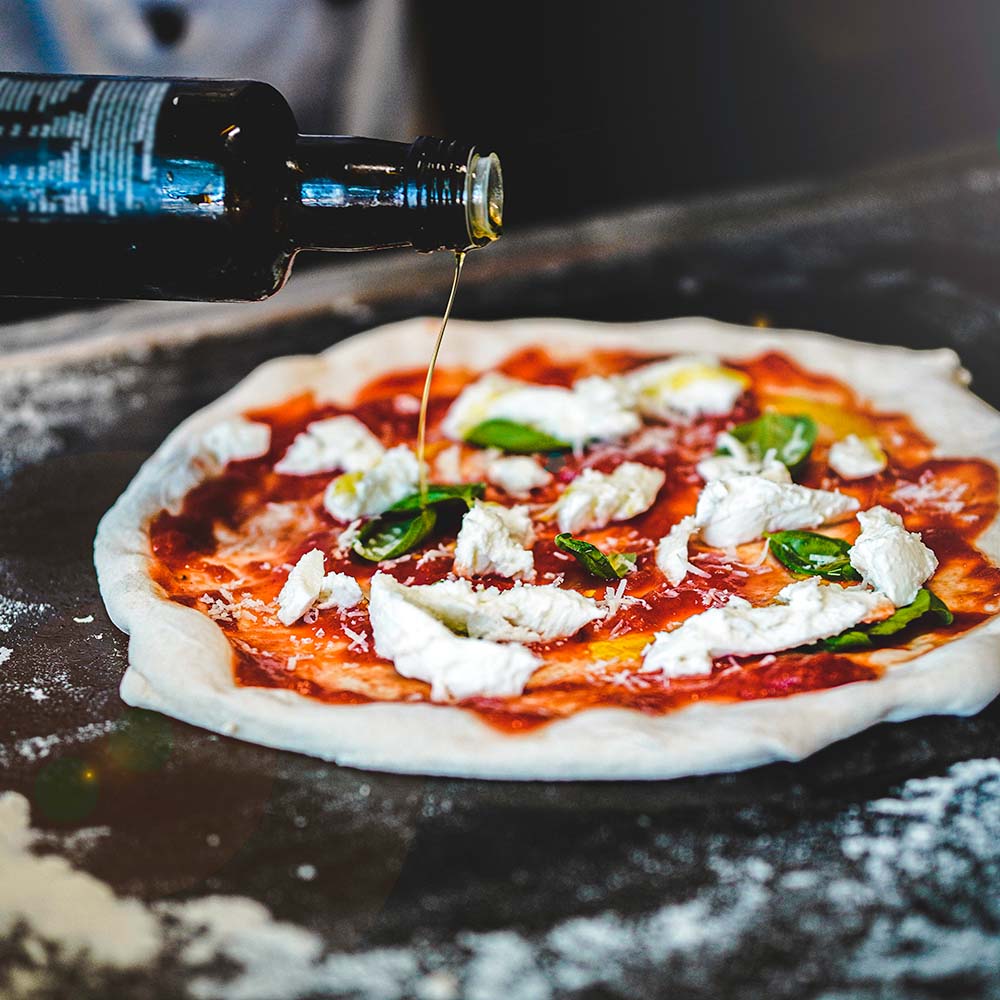 En rå pizza bund ligger på en melstøvet overflade, toppet med tomatsauce, klatter af hvid mozzarella og friske basilikumblade. En flaske med olivenolie hældes over pizzaen.