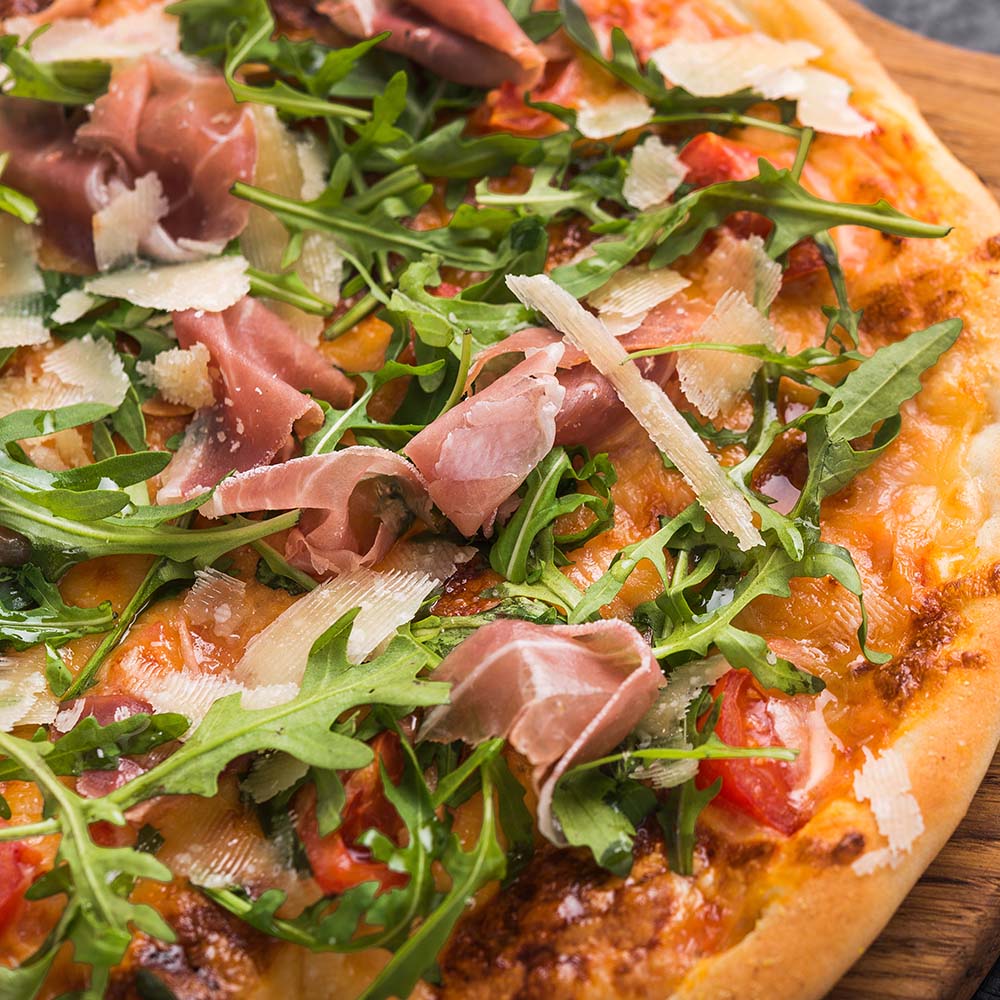 En pizza med en sprød bund dækket af frisk rucola, skiver af prosciutto og let smuldret parmesanost.
